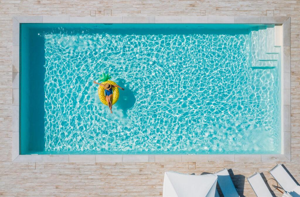 Frau im Bikini, mit Strohhut und einer Sonnenbrille, die im Pool auf riesiger aufblasbarer gelber Ananas schwimmt