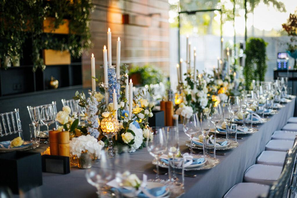 festlich gedeckte Tafel mit grauem Tischtuch, transparenten Stühlen, silbernen Tellern, weißen und blauen Blumen und Kerzen